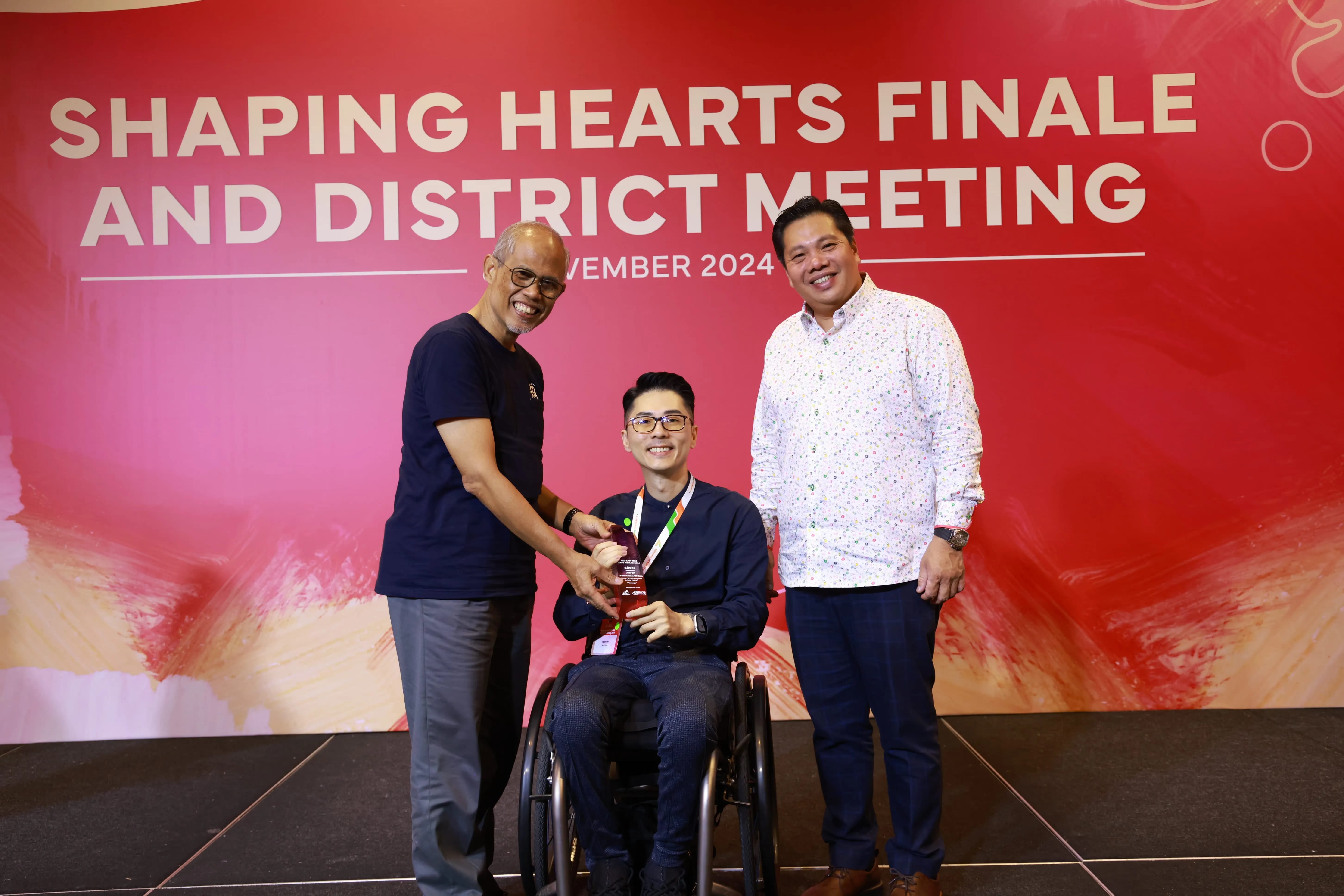 Man in wheelchair with two men standing on both sides in front of a backdrop that reads "Shaping Hearts Finale and District Meeting November 2024."